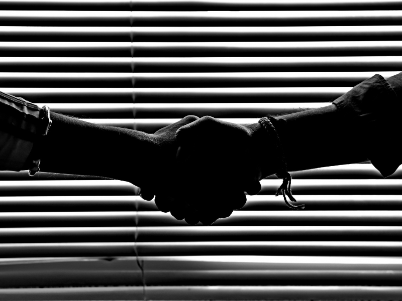 Black and white photo of 2 hands shaking in front of window blinds. The blinds cast a shadow so that the hands appear black against a black/white striped background.